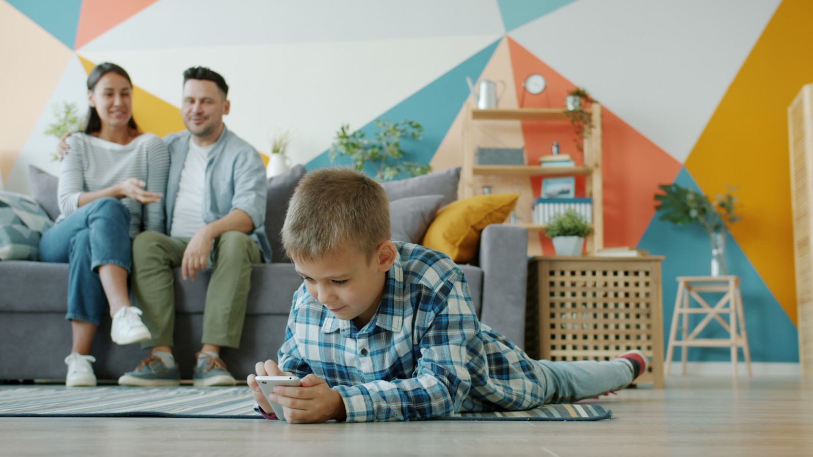 Boy plays on phone while parents watch from couch.